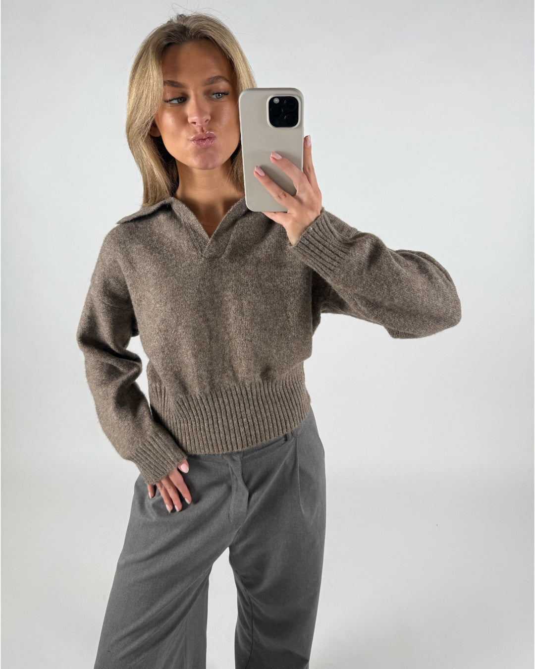 Person tager en selfie iført en brun sweater og grå bukser, poserende i en neutral indendørs indstilling.