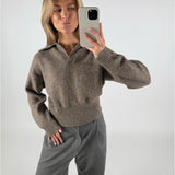 Person tager en selfie iført en brun sweater og grå bukser, poserende i en neutral indendørs indstilling.