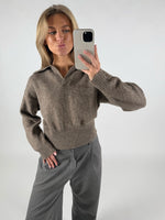 Person tager en selfie iført en brun sweater og grå bukser, poserende i en neutral indendørs indstilling.