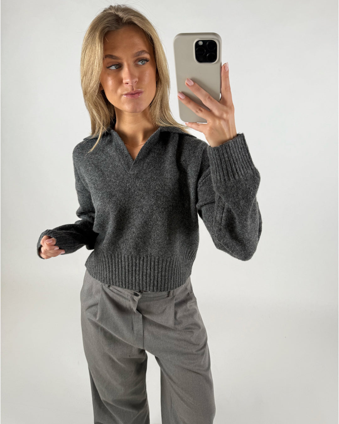 Person holder en mobiltelefon og tager et spejlfoto iført en grå sweater og bukser, stående mod en lys baggrund.