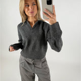 Person holder en mobiltelefon og tager et spejlfoto iført en grå sweater og bukser, stående mod en lys baggrund.