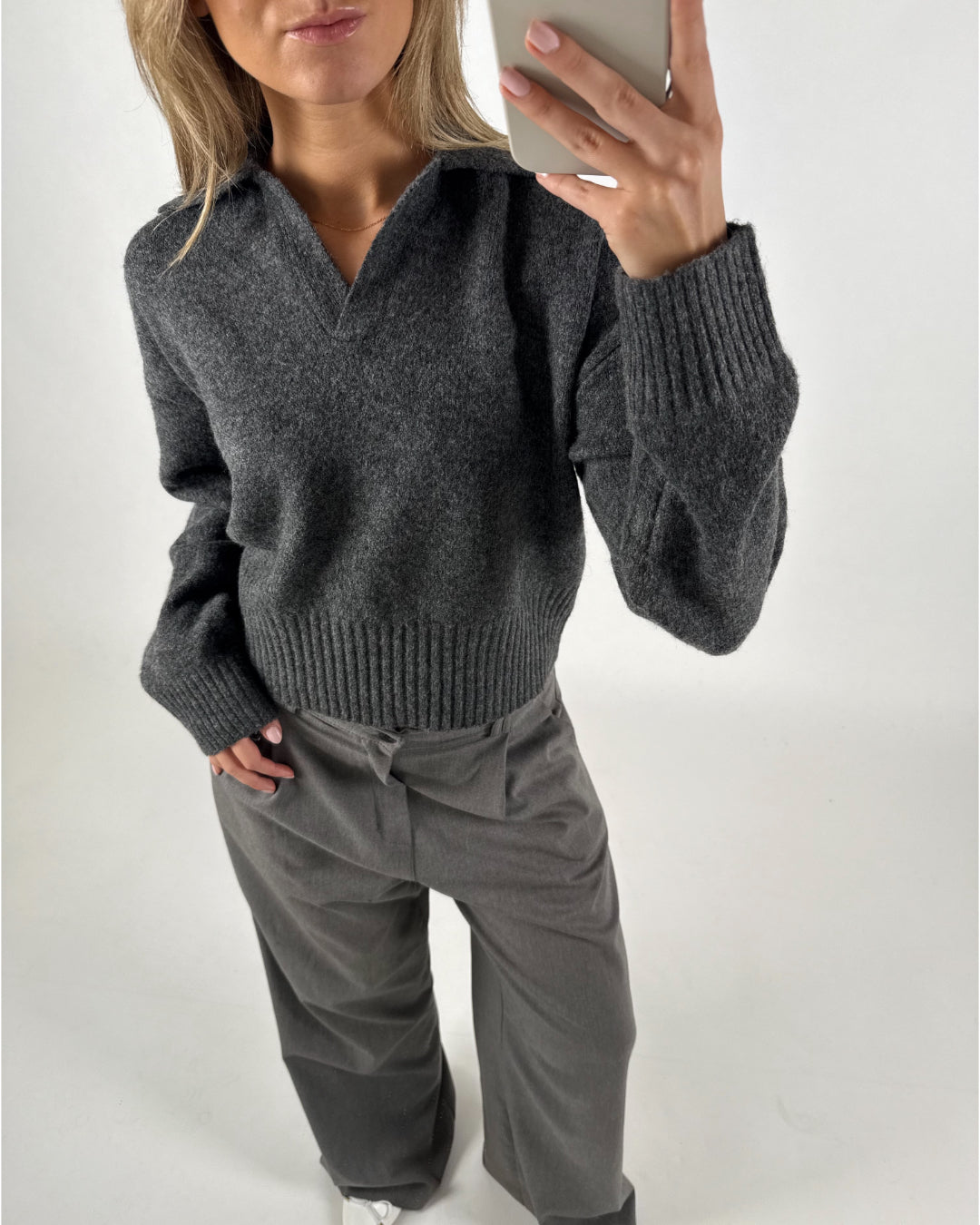 En person iført en mørkegrå trøje og lysegrå bukser tager en selfie i et lyst rum, mens de står oprejst med en hånd i lommen og kigger i spejlet.