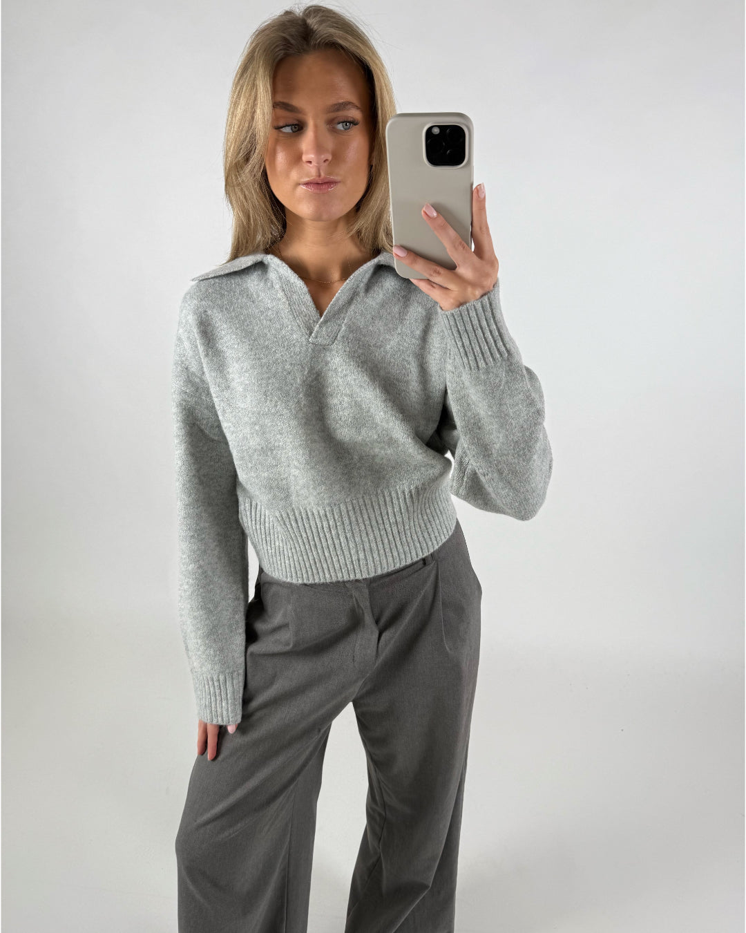 En person tager en selfie iført en lys grå trøje og mørke bukser. Personen står i et enkelt rum med en lys baggrund.