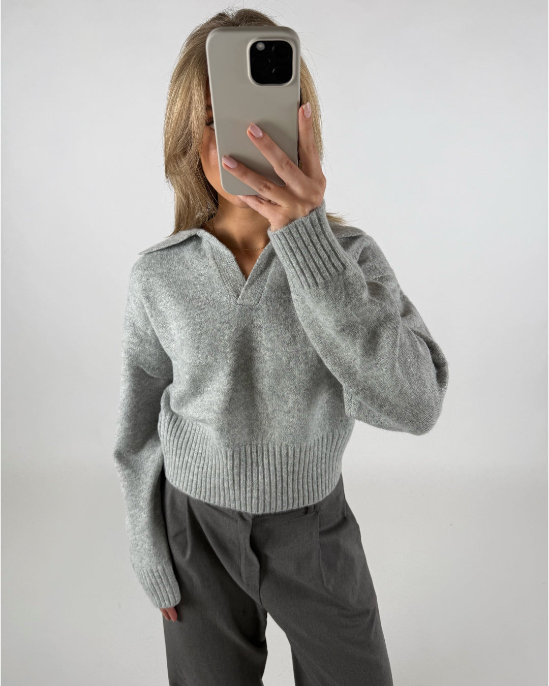 Person tager et spejlbillede med en mobiltelefon, iført en grå sweater med krave og mørke bukser. Baggrunden er neutral og lysegrå.