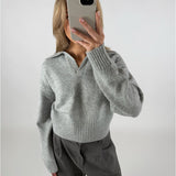 Person tager et spejlbillede med en mobiltelefon, iført en grå sweater med krave og mørke bukser. Baggrunden er neutral og lysegrå.