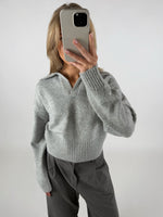 Person tager et spejlbillede med en mobiltelefon, iført en grå sweater med krave og mørke bukser. Baggrunden er neutral og lysegrå.