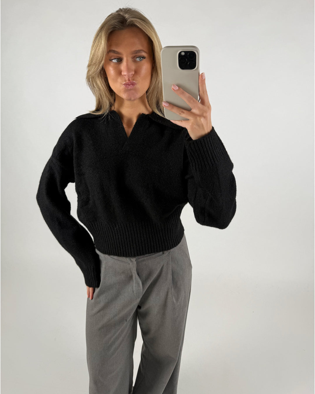 Kvinde tager en selfie i en spejlglat rum, iført sort sweater og grå bukser, med løst hår og laver en kysmund.