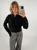 Kvinde tager en selfie i en spejlglat rum, iført sort sweater og grå bukser, med løst hår og laver en kysmund.