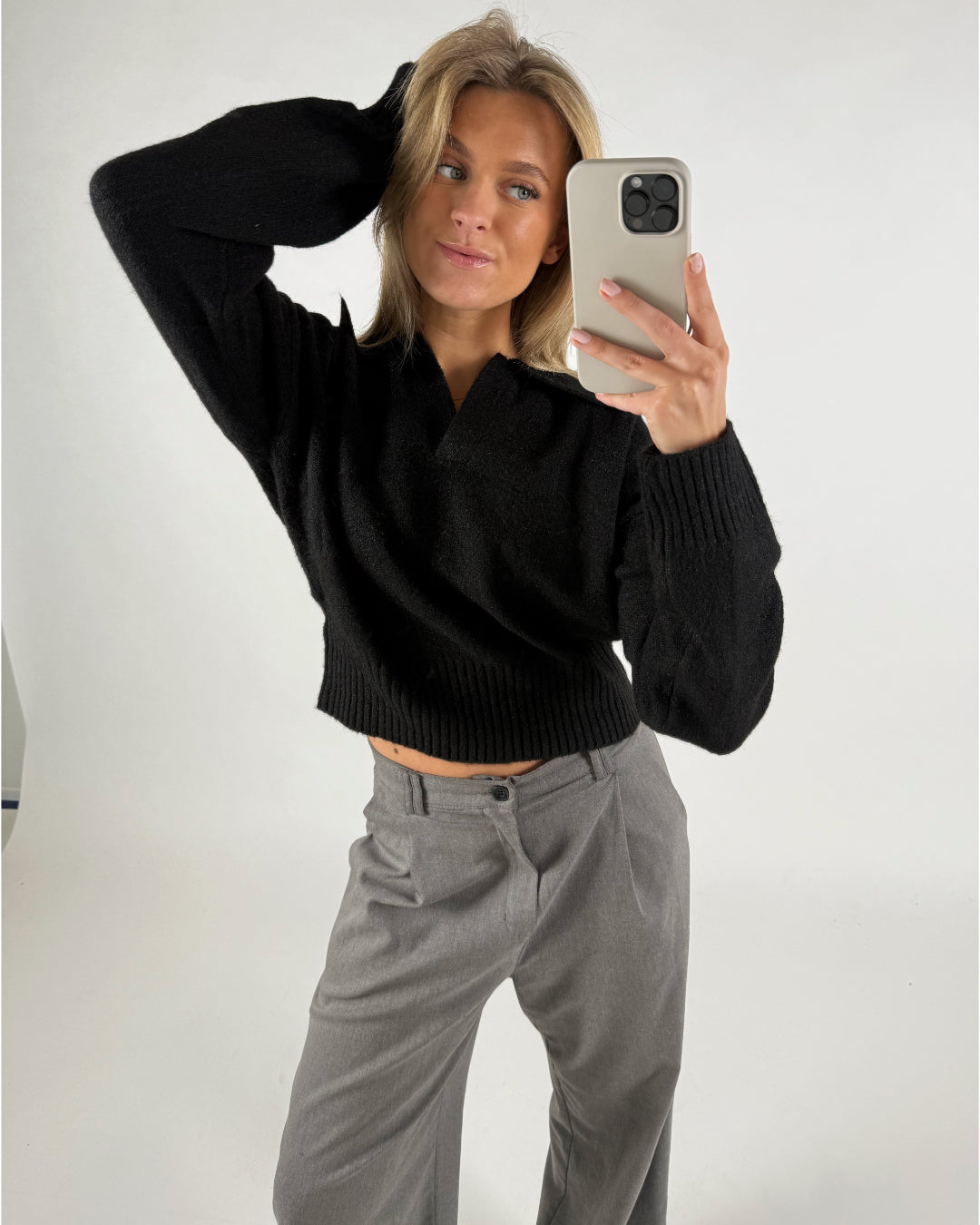 En person tager en selfie iført en sort sweater og grå bukser, poserer med hånden på hovedet i et neutralt belyst rum.