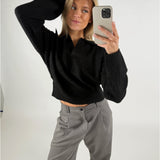 En person tager en selfie iført en sort sweater og grå bukser, poserer med hånden på hovedet i et neutralt belyst rum.