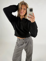 En person tager en selfie iført en sort sweater og grå bukser, poserer med hånden på hovedet i et neutralt belyst rum.