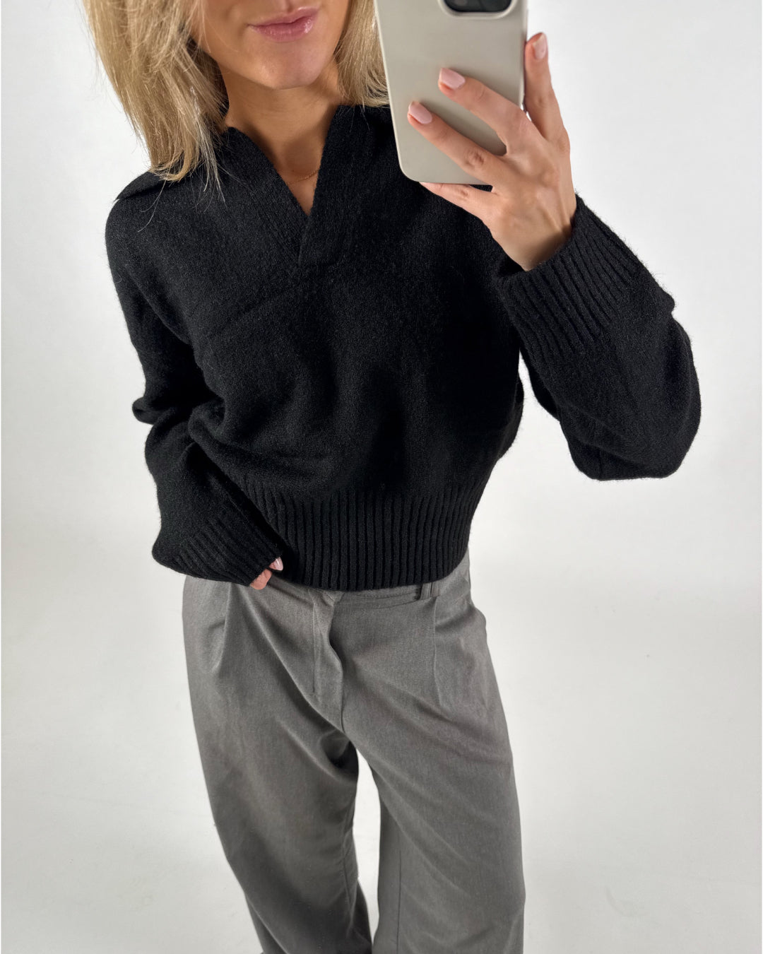 Person tager et billede med en mobiltelefon. De har en sort sweater og grå bukser på. Baggrunden er simpel og hvid.