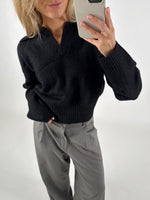 Person tager et billede med en mobiltelefon. De har en sort sweater og grå bukser på. Baggrunden er simpel og hvid.