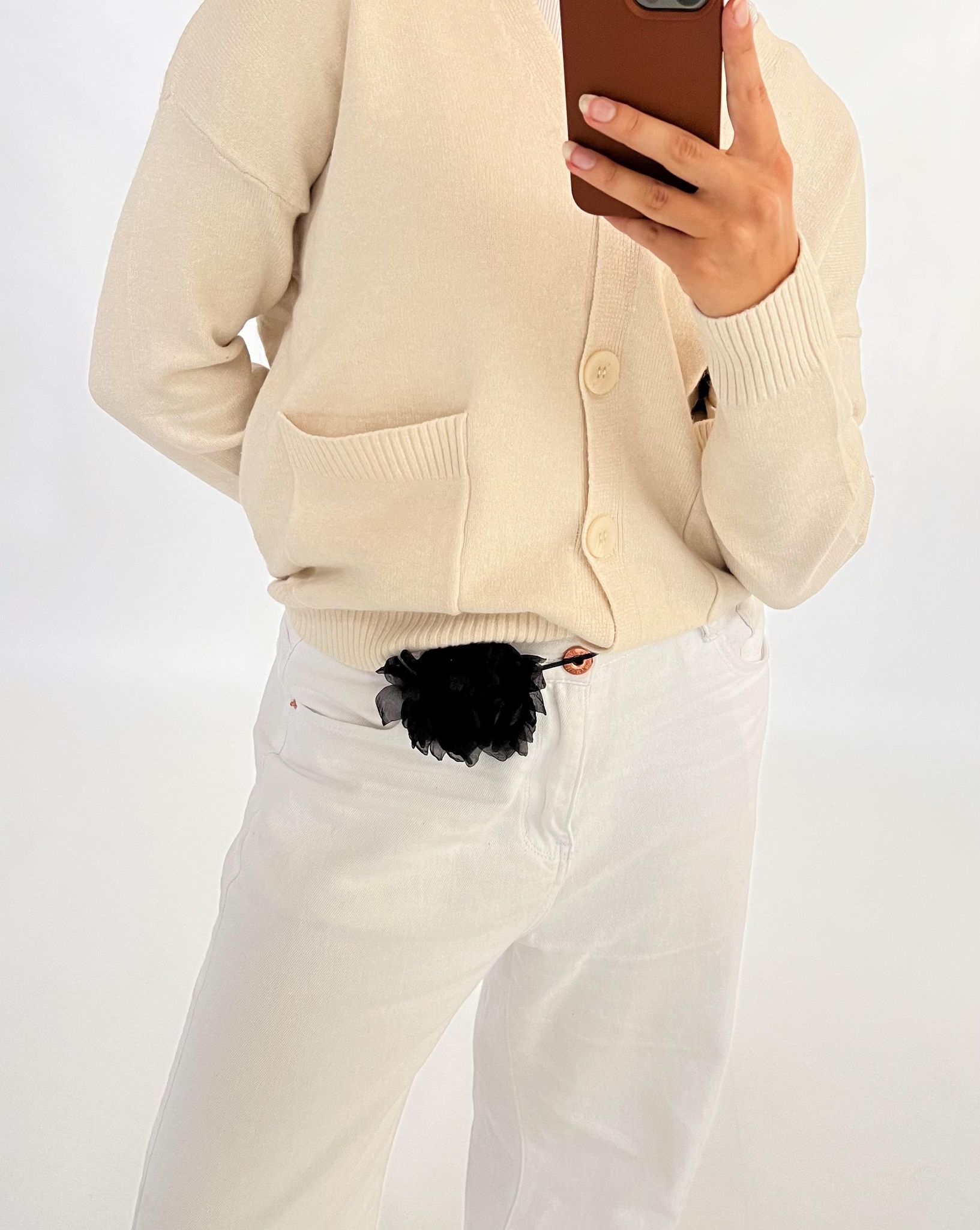 Person iført en lys sweater tager et selfie med en brun mobiltelefon. De har hvide bukser med en sort blomst ved bæltet, stående i et lyst, neutralt rum.