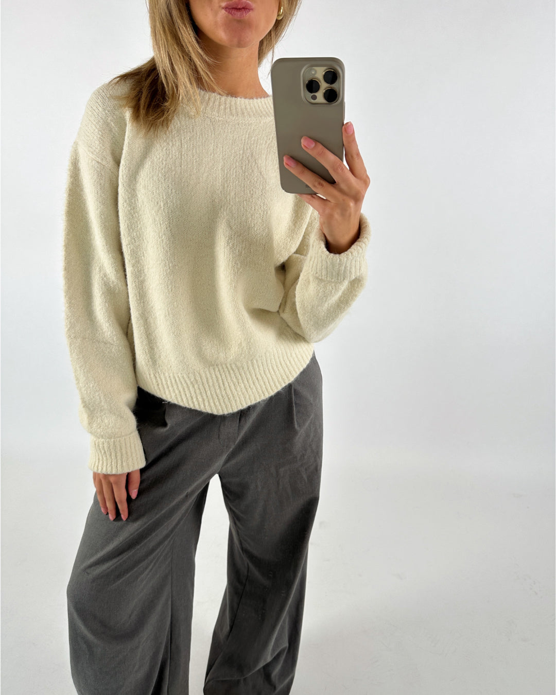 Person i cremefarvet sweater tager et billede med en telefon. De har mørke bukser på og poserer i et neutralt rum.