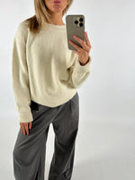 Person i cremefarvet sweater tager et billede med en telefon. De har mørke bukser på og poserer i et neutralt rum.