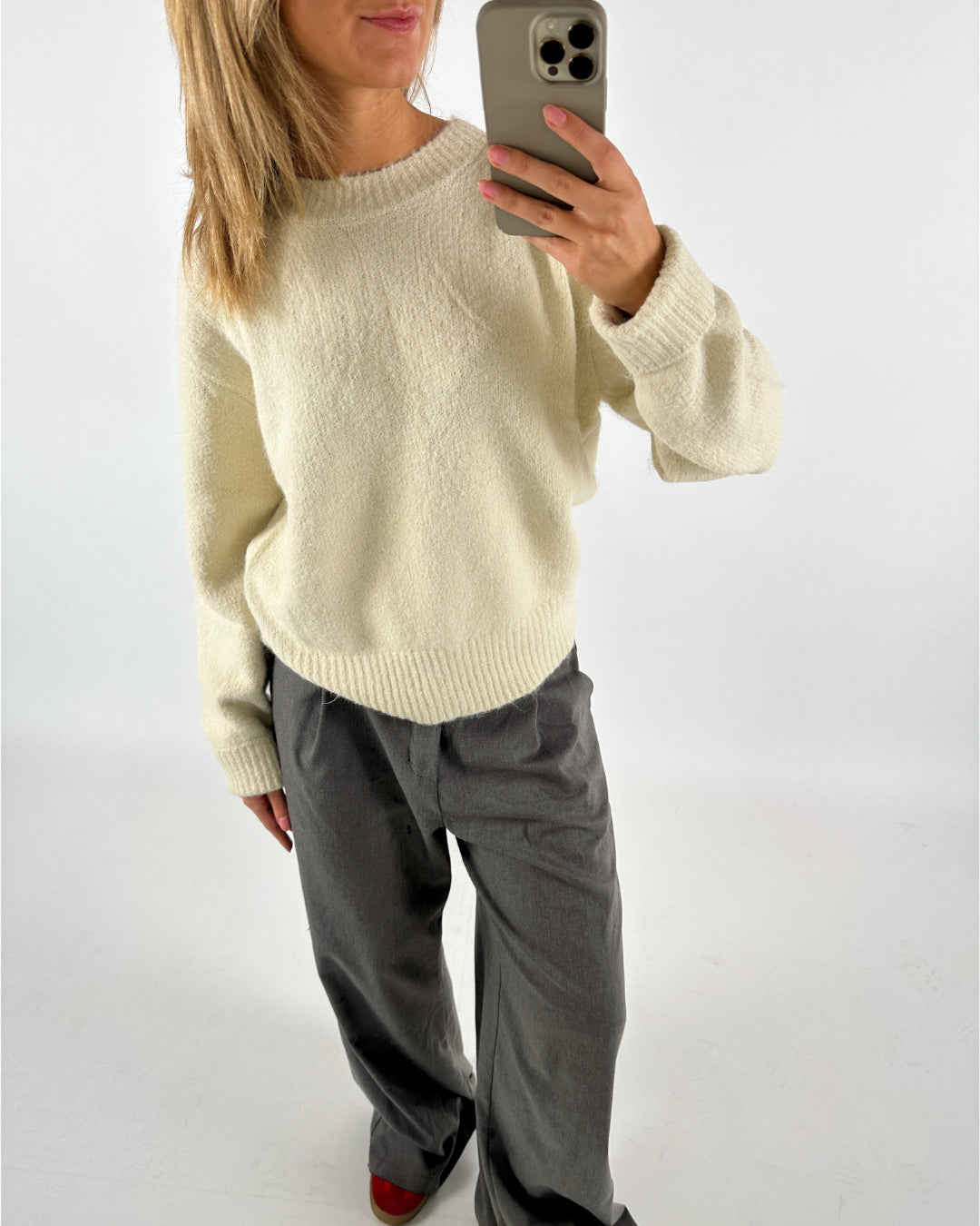 Kvinde iført cremefarvet sweater og grå bukser tager en selfie med en smartphone i oplyst indendørs miljø.
