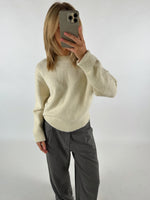 Person tager et spejlbillede, iført en cremefarvet sweater og grå bukser, i et hvidt rum. Personen holder en mobiltelefon op til ansigtet og har skulderlangt hår.