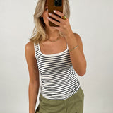 En person tager en selfie med en mobiltelefon, iført en sort/hvid-stribet tanktop og grønne bukser, mod en neutral baggrund.
