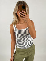 En person tager en selfie med en mobiltelefon, iført en sort/hvid-stribet tanktop og grønne bukser, mod en neutral baggrund.