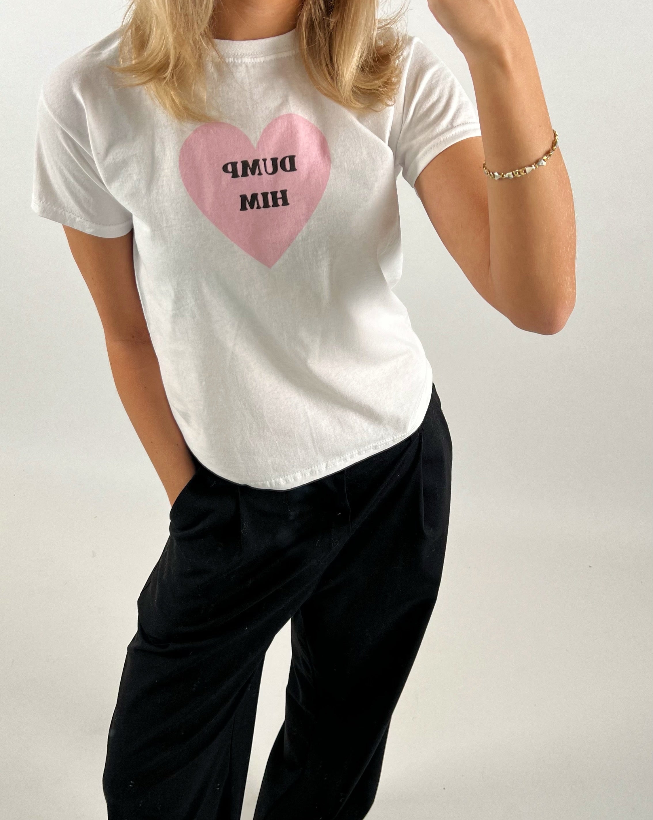 En person iført en hvid T-shirt med en lyserød hjertegrafik, der viser teksten "DUMP HIM", står afslappet med den ene hånd i lommen i et neutralt rum.