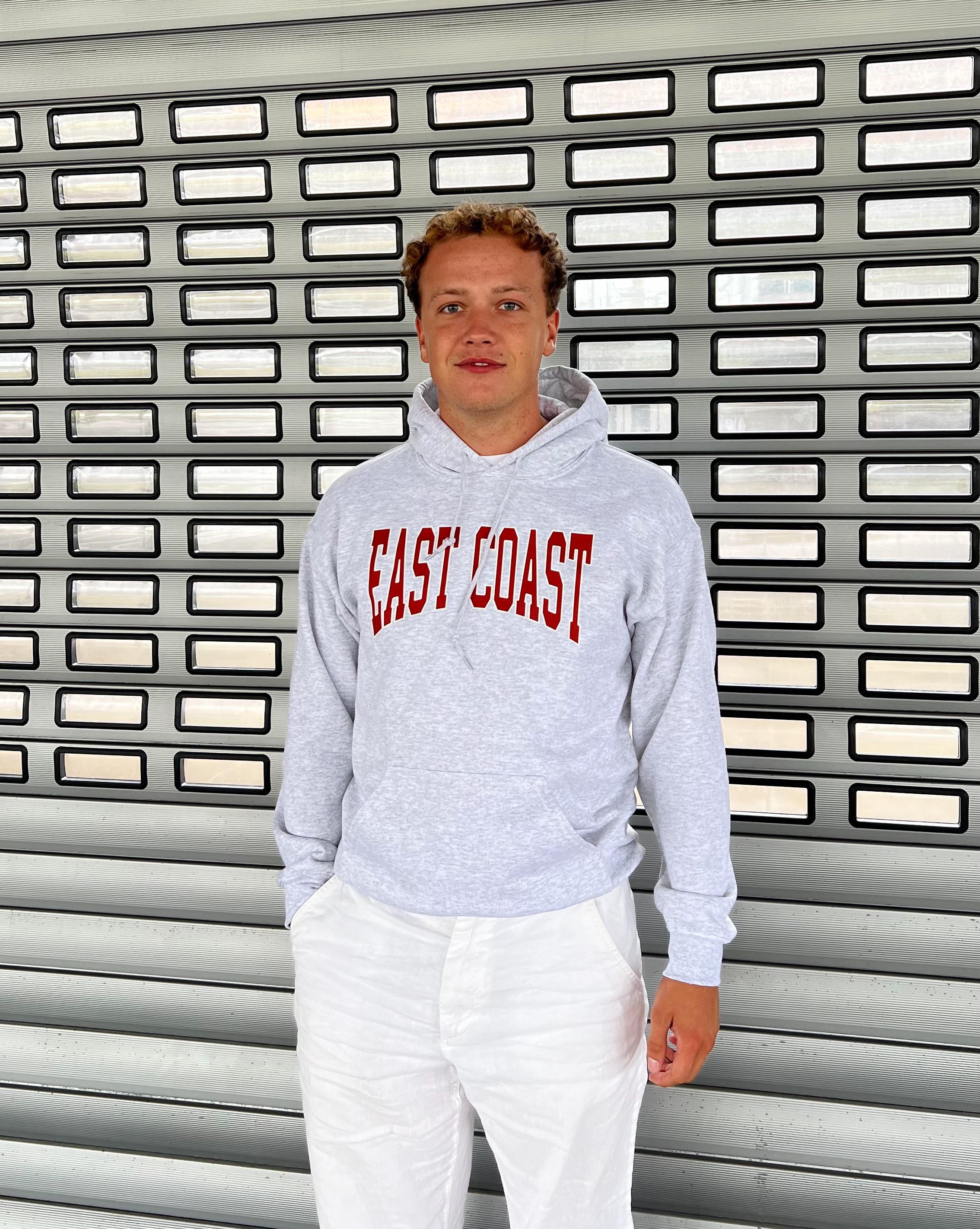 En person står foran en metaldør iført en grå hættetrøje med påskriften "EAST COAST" i rødt og hvide bukser. Baggrunden består af små rektangulære vinduer.