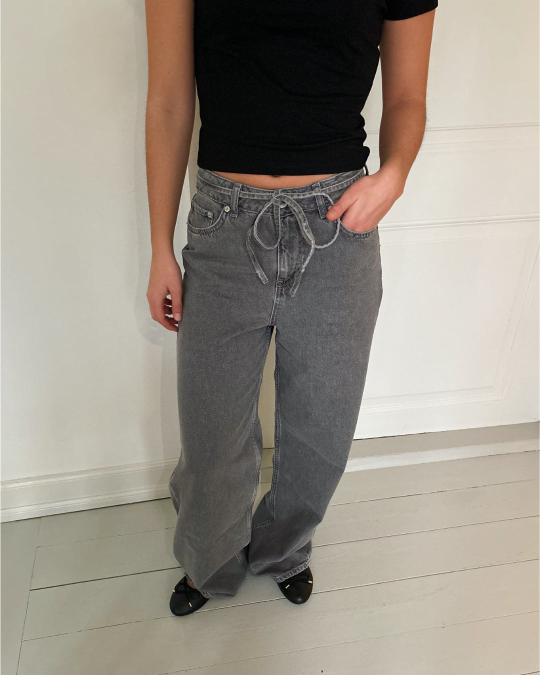 En person iført grå jeans med snøre og sort trøje står på et lyst trægulv foran en hvid væg.