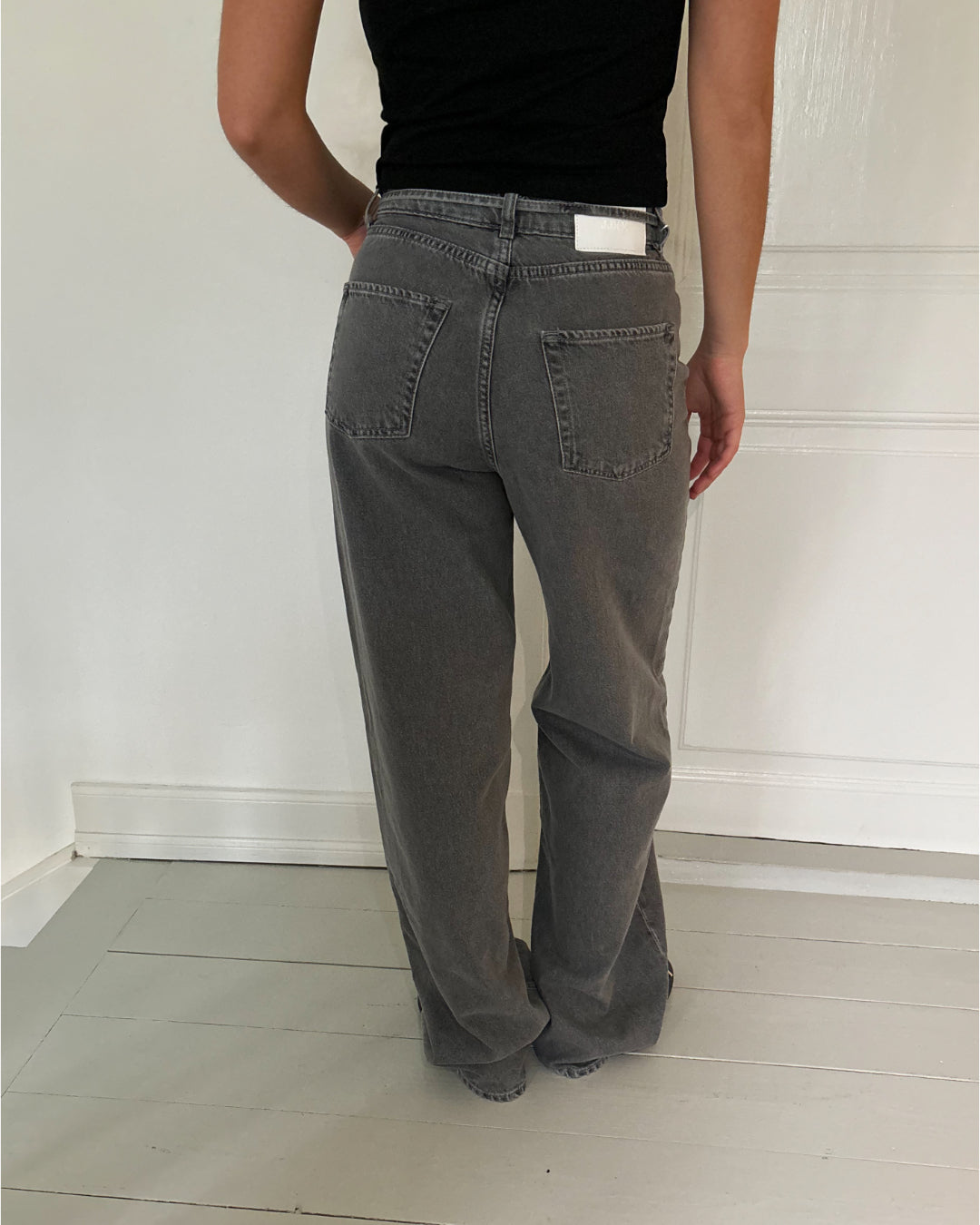 En person iført grå jeans og sort top står med ryggen til i et rum med lyse trægulve og en hvid væg i baggrunden.
