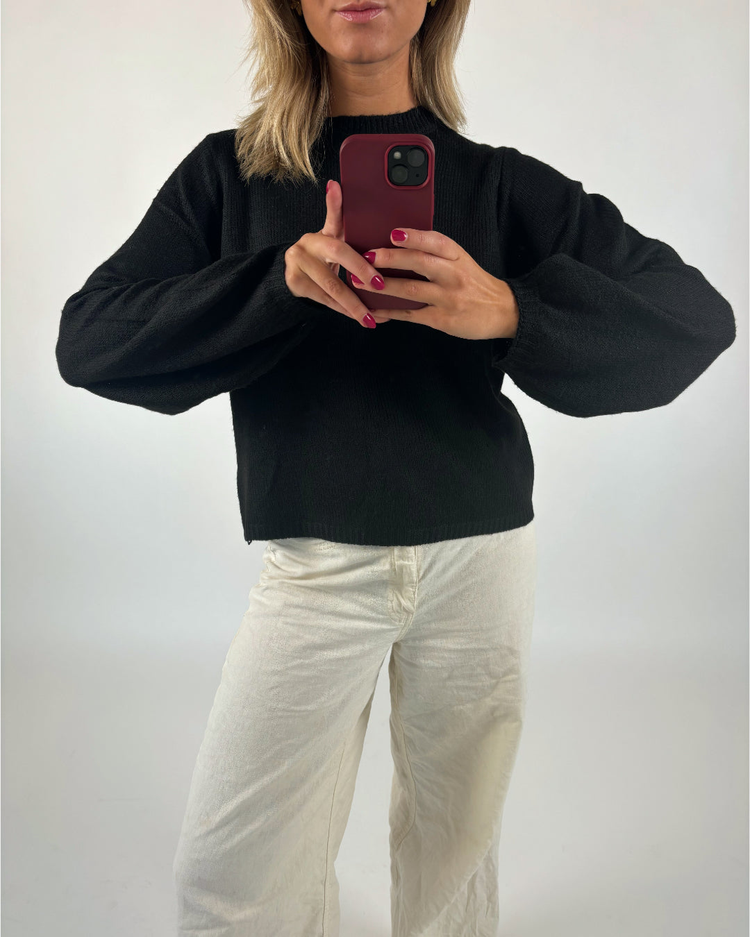 Person tager selfie med rød mobiltelefon iført sort trøje og hvide bukser; står i et rum med hvid baggrund.