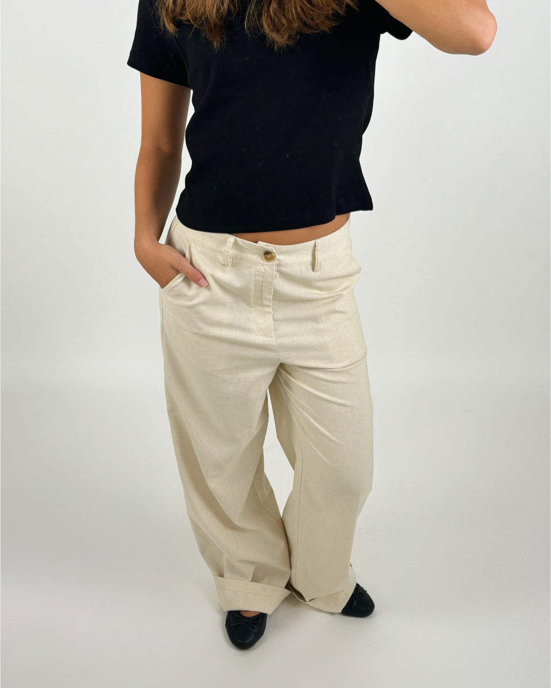 En person i sort t-shirt og beige bukser står med hånden i lommen. Personen har sorte sko på, og baggrunden er en ensfarvet væg.