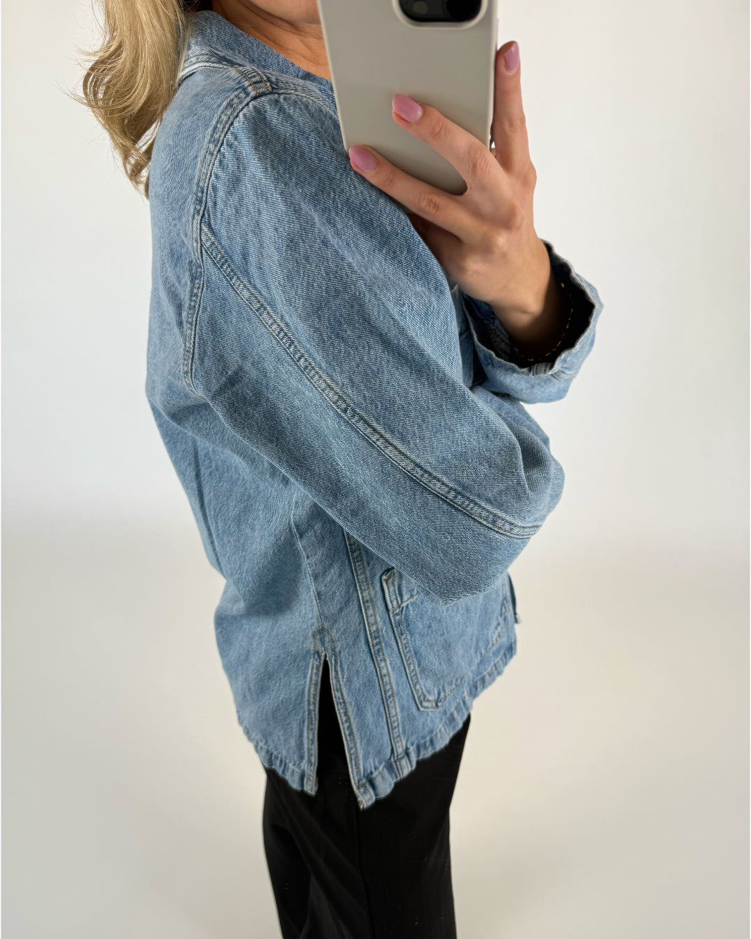 En person iført en denimjakke tager et selfie med en smartphone. Personen står i en enkeltfarvet, lys baggrund og har sort tøj på under jakken.