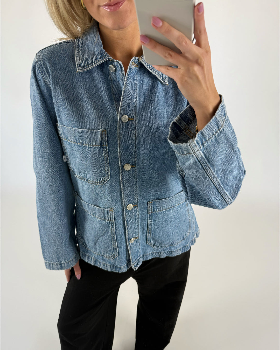 En person iført en blå denimjakke tager en selfie i et lyst rum. Jakken har store lommer og knapper foran, og personen har lyserøde negle.