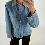 En person iført en blå denimjakke tager en selfie i et lyst rum. Jakken har store lommer og knapper foran, og personen har lyserøde negle.