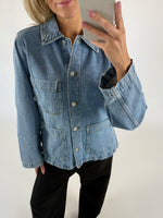En person iført en blå denimjakke tager en selfie i et lyst rum. Jakken har store lommer og knapper foran, og personen har lyserøde negle.