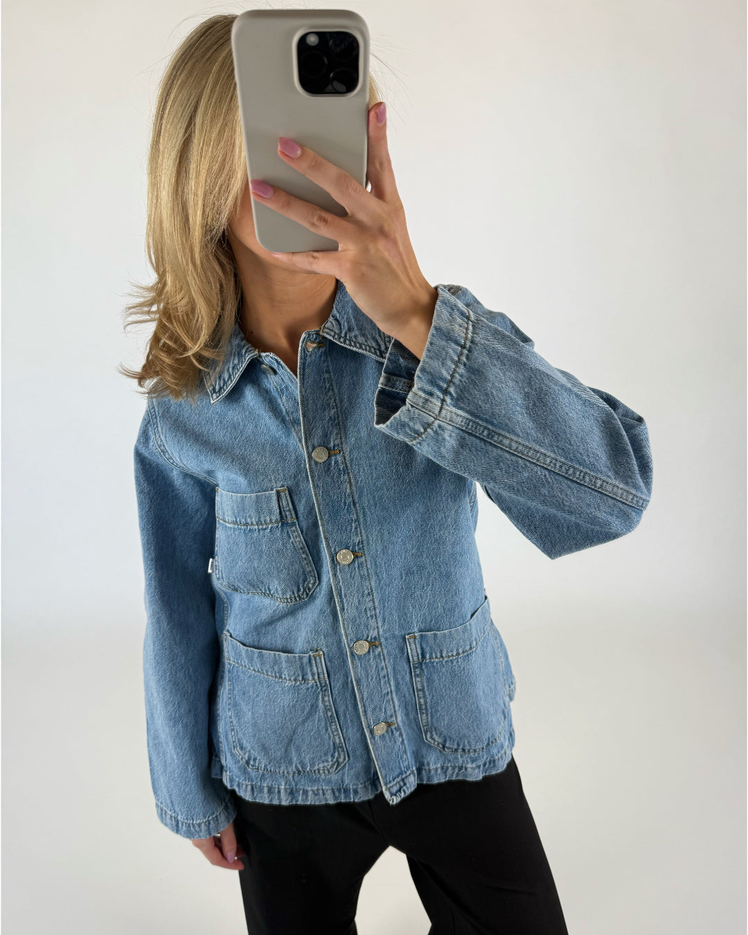 En person iført en blå denimjakke tager en selfie med en smartphone i et neutralt belyst rum. Jakken har store frontlommer og metalknapper.