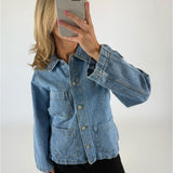 En person iført en blå denimjakke tager en selfie med en smartphone i et neutralt belyst rum. Jakken har store frontlommer og metalknapper.