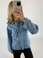 En person iført en blå denimjakke tager en selfie med en smartphone i et neutralt belyst rum. Jakken har store frontlommer og metalknapper.
