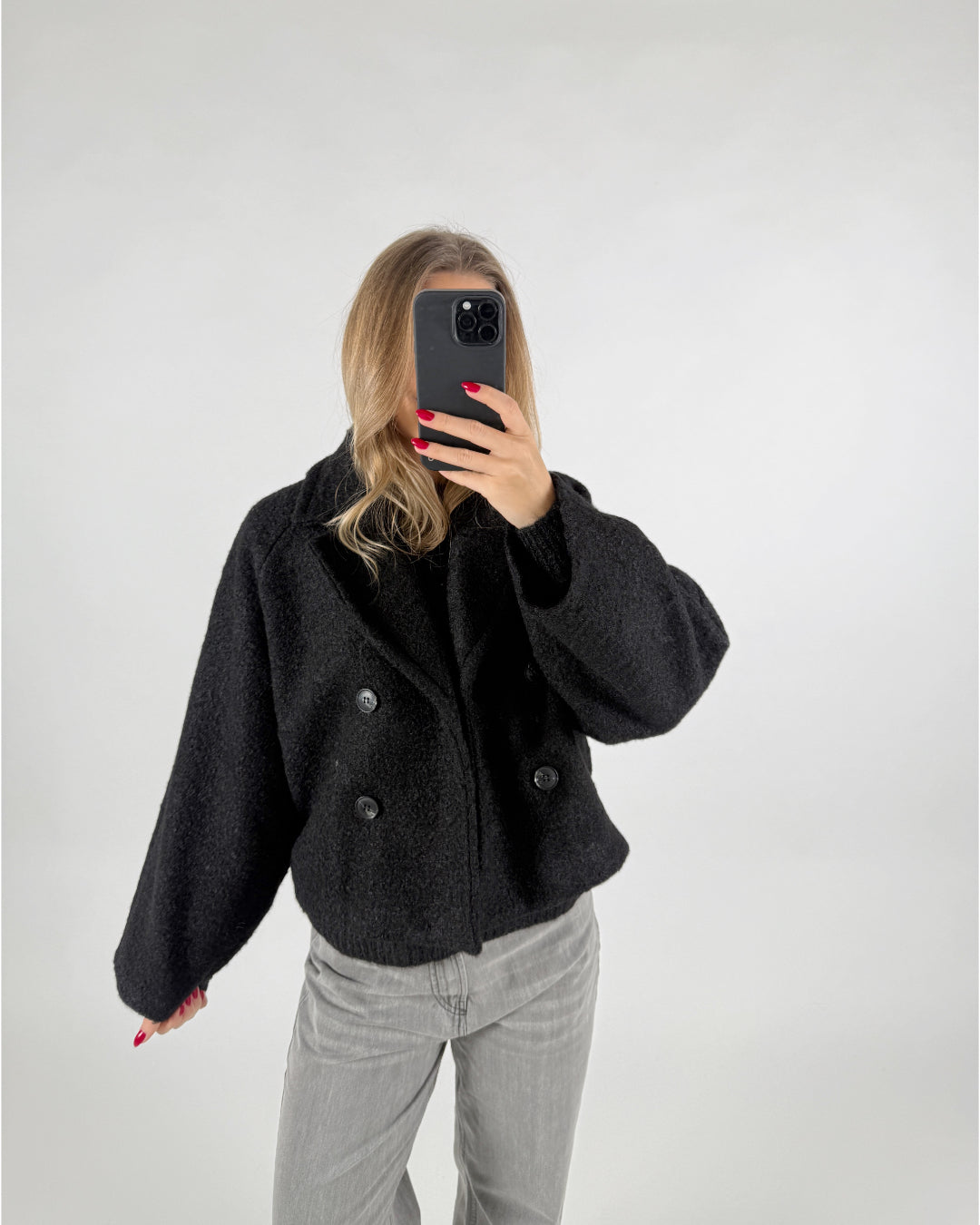 Person tager et billede af sig selv, iført en sort jakke med dobbeltknap, i et lyst rum. Personen holder en mobiltelefon op foran ansigtet med rødmalede negle.