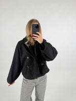 Person tager et billede af sig selv, iført en sort jakke med dobbeltknap, i et lyst rum. Personen holder en mobiltelefon op foran ansigtet med rødmalede negle.