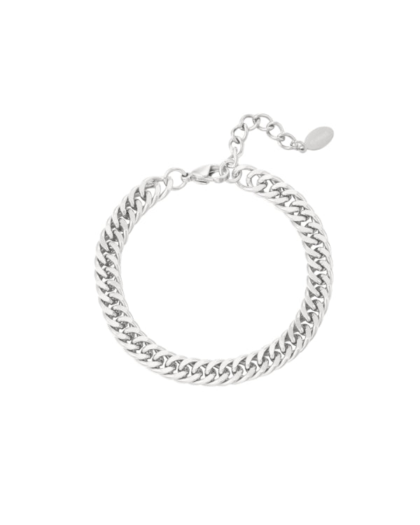 Armbånd i sølvfarvet metallisk materiale ligger i en cirkel. Det har en kædelignende struktur med en justerbar lukning og en lille vedhæng. Baggrunden er hvid og ren.