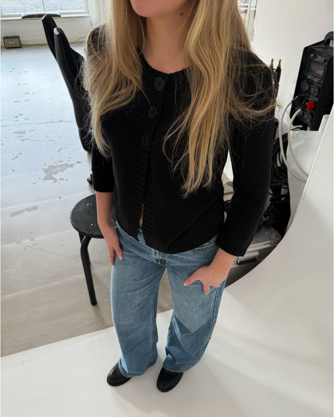 En person med langt blondt hår, iført en sort cardigan og blå jeans, står i et lyst rum ved siden af en stol og udstyr.