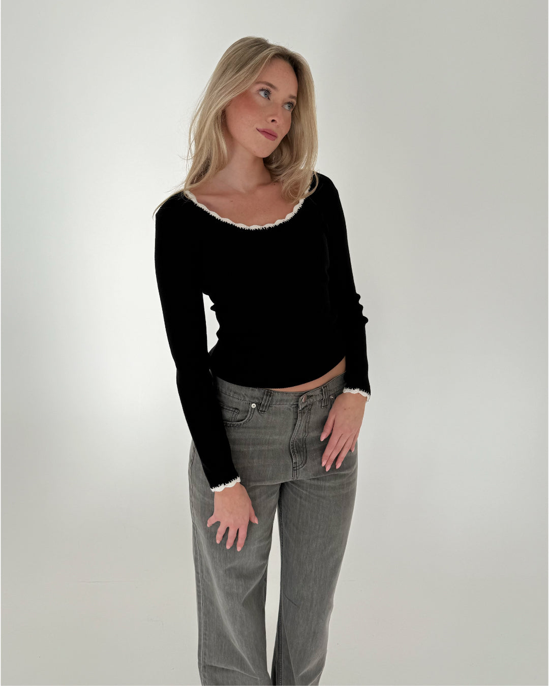 En person iført en sort trøje og grå jeans står afslappet i en minimalistisk, hvidt belyst baggrund.