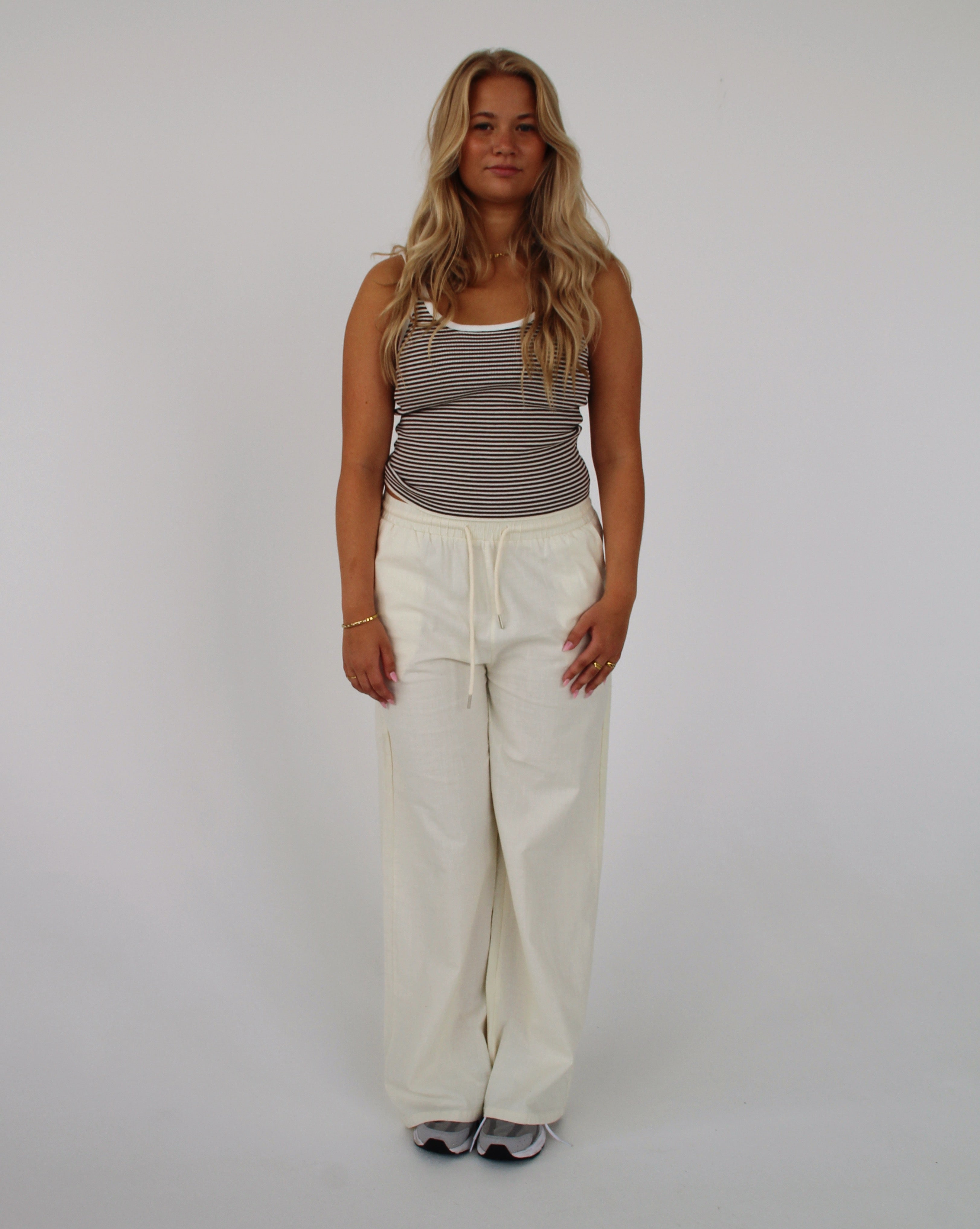En person står stille iført en stribet tanktop og hvide bukser. Personen er i et lyst rum med en ensfarvet baggrund.