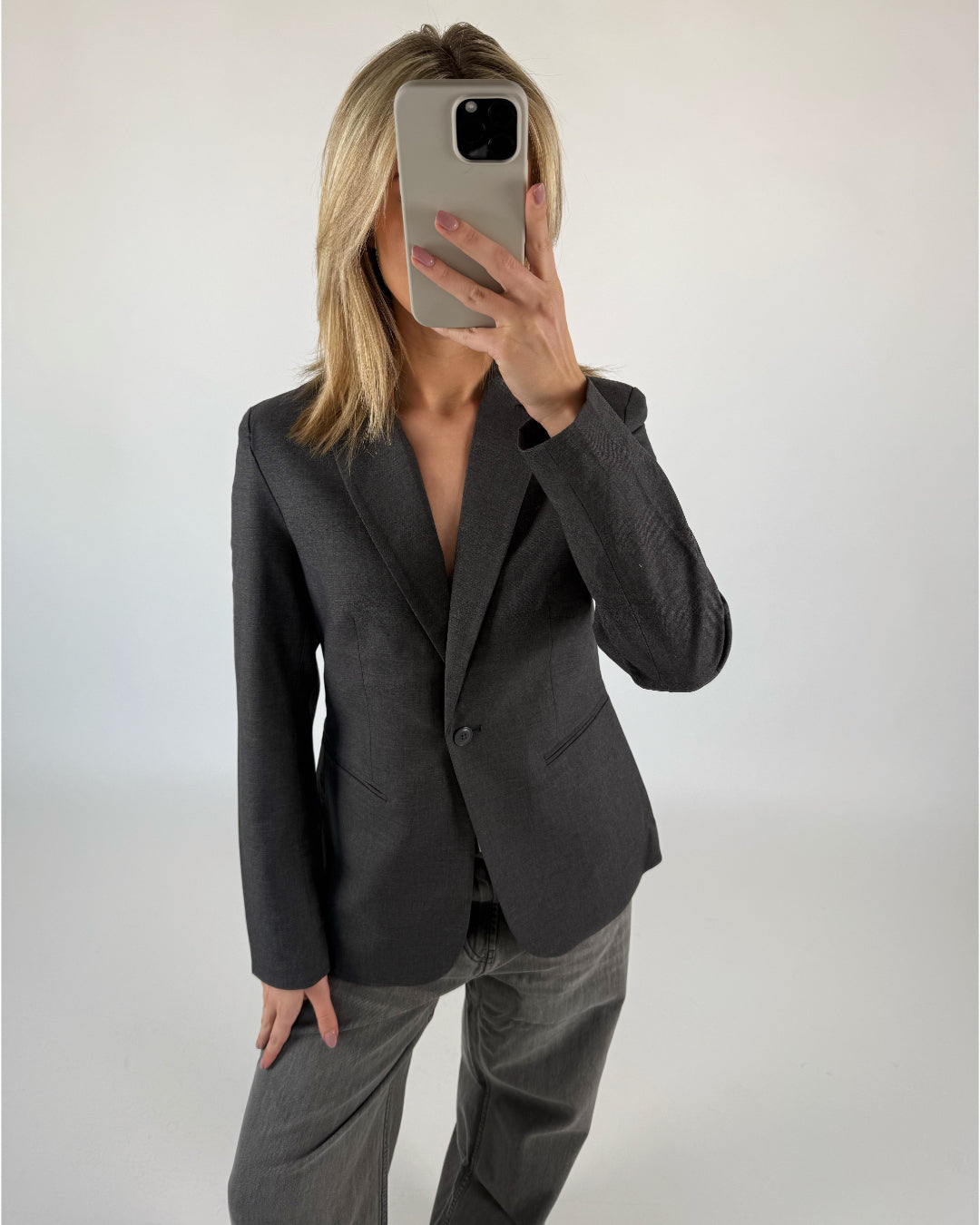 Person iført mørk blazer tager selfie med mobiltelefon, ansigt dækket af telefonen, i et neutralt, lyst rum.