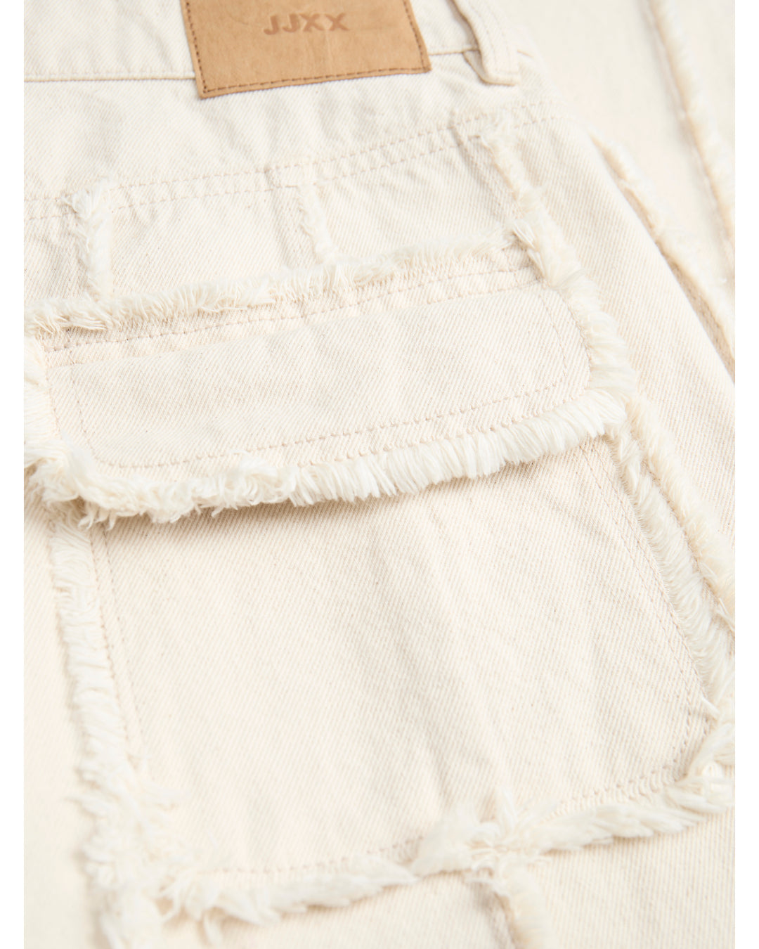 En beige denimjakke med frynset detalje på lommen. Tekst på en brun etiket viser "JJXX". Jakken ligger fladt i en lys kontekst.
