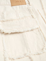En beige denimjakke med frynset detalje på lommen. Tekst på en brun etiket viser "JJXX". Jakken ligger fladt i en lys kontekst.