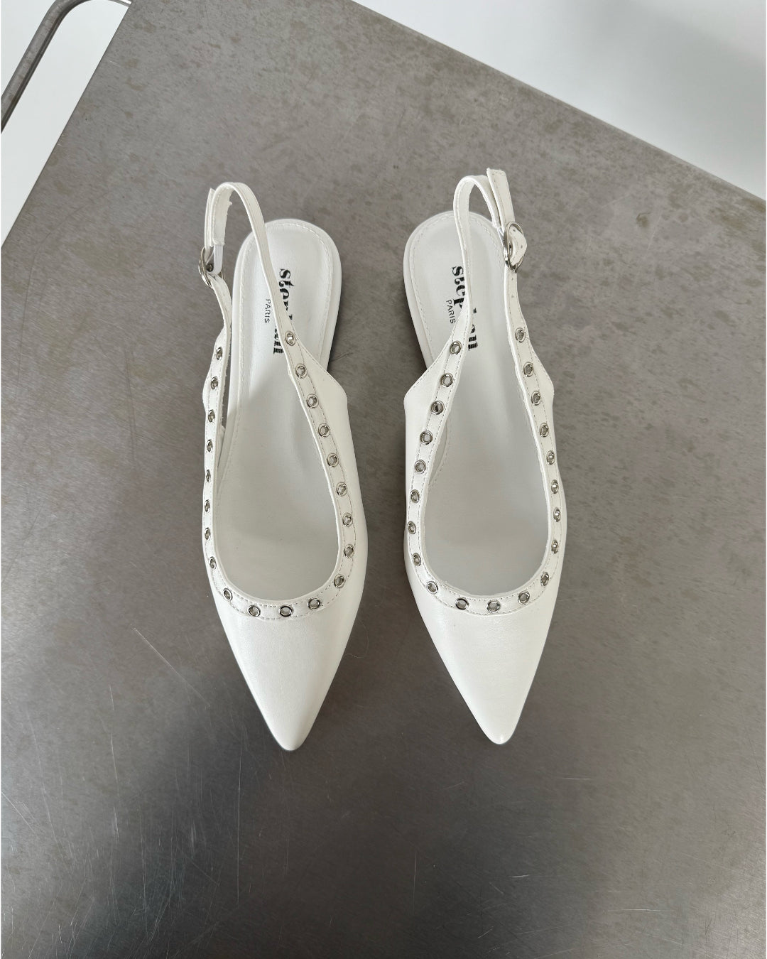 Hvide slingback-sko med spidse snuder dekoreret med sølvnitter, står på en metallisk overflade. Skoens indersål har teksten "stevemai PARIS".