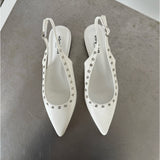 Hvide slingback-sko med spidse snuder dekoreret med sølvnitter, står på en metallisk overflade. Skoens indersål har teksten "stevemai PARIS".