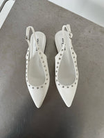 Hvide slingback-sko med spidse snuder dekoreret med sølvnitter, står på en metallisk overflade. Skoens indersål har teksten "stevemai PARIS".