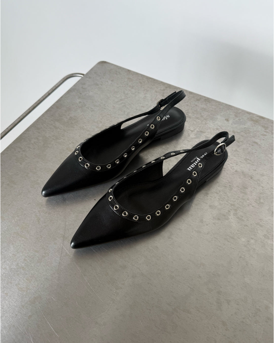 Sorte spidse sko med nitter ligger på et metalbord. Tekst på skoen læser: "PRADA". Minimalistisk og lys baggrund uden andre genstande.